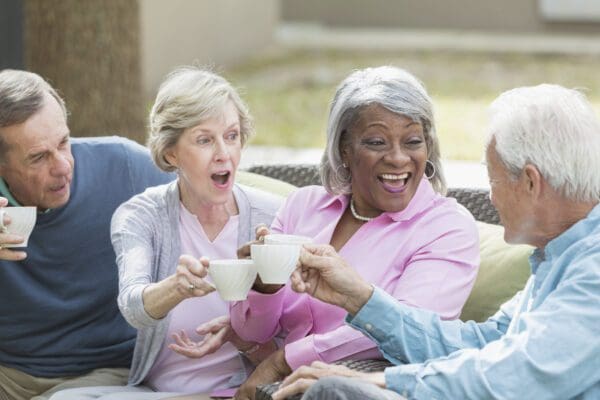 A group of four multi-ethnic seniors sitting together on patio furniture outdoors, talking and smiling, drinking tea or coffee. One of the men and the women are toasting with their cups, excited expressions on their faces. They are in retirement, enjoying spending time with friends.
